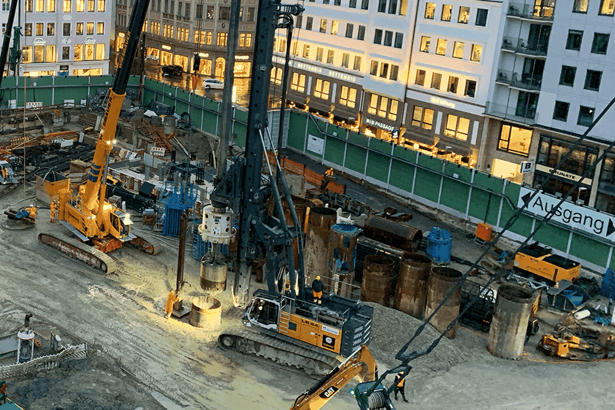 A construction site with machines and workers, surrounded by buildings and a security fence.
