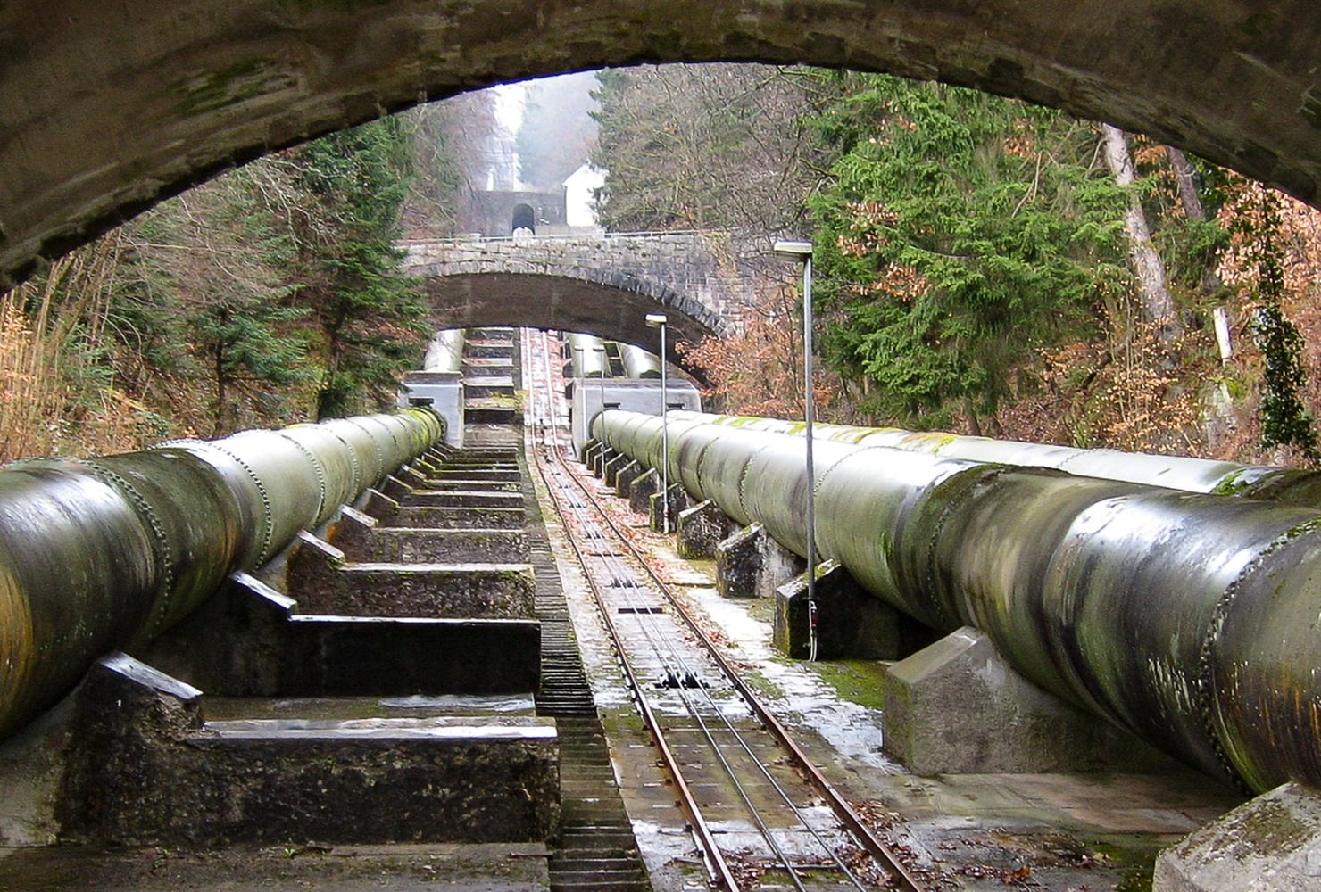View of large industrial pipes running under several stone arch bridges, surrounded by trees and moss-covered structures.