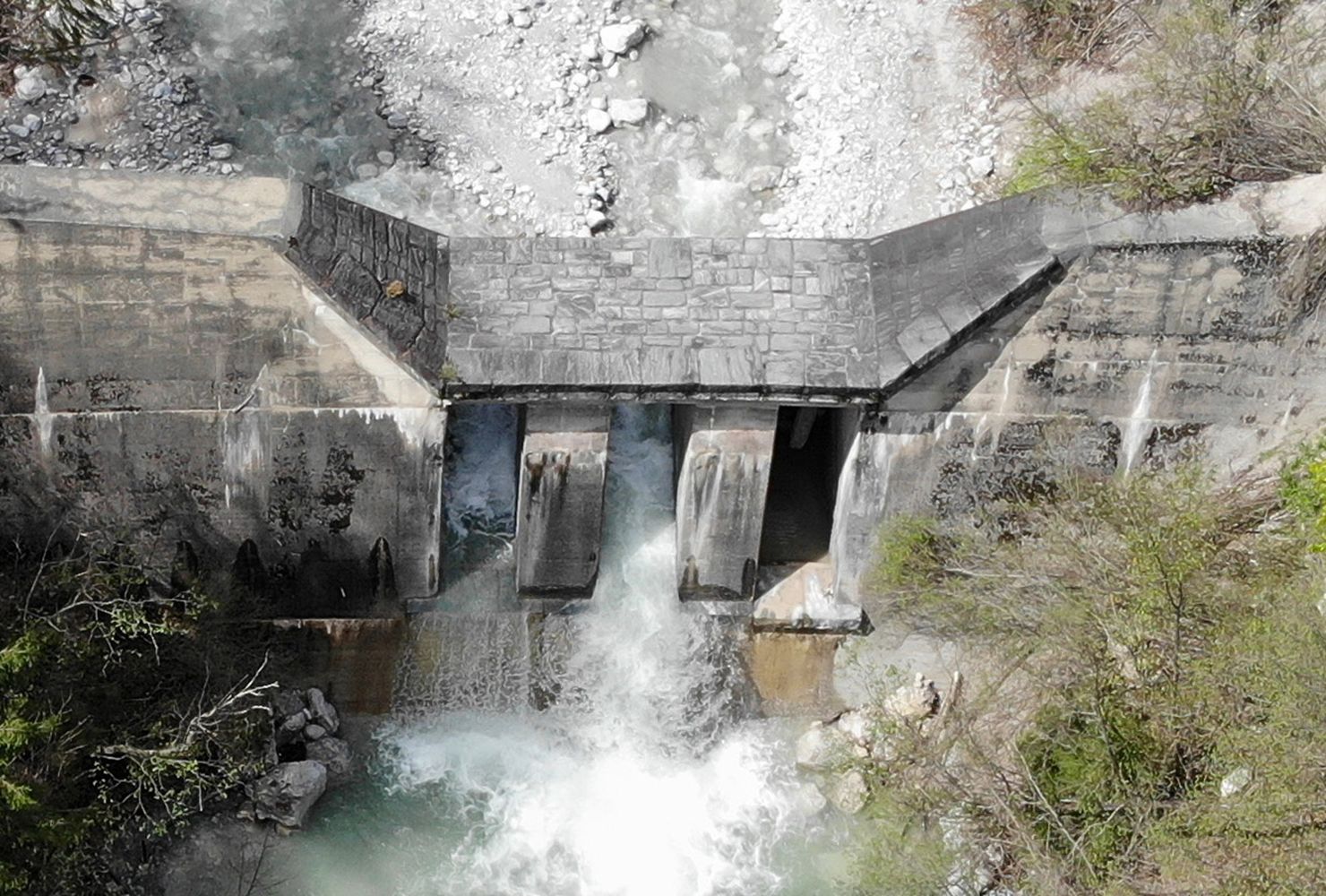 Aerial view of a concrete dam with water flowing through three openings, surrounded by lush greenery and rocky terrain.