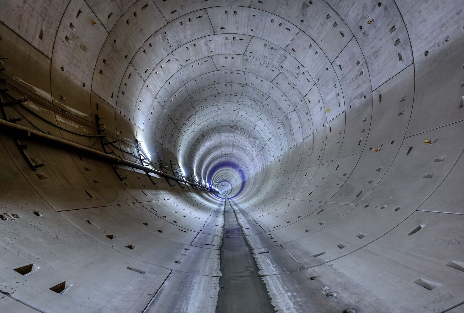 View inside a long, cylindrical concrete tunnel, with evenly spaced panels and cables running along the left side, creating a vanishing point.