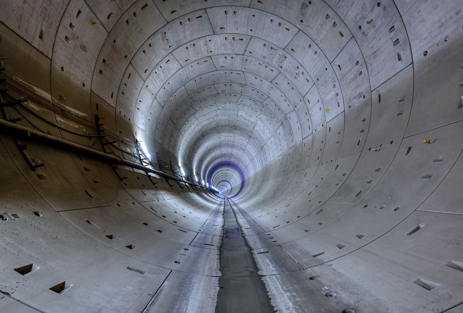 Innenansicht eines großen, kreisförmigen Betontunnels mit glattem Boden, der sich in die Ferne erstreckt und an der linken Wand von Kabeln gesäumt ist.