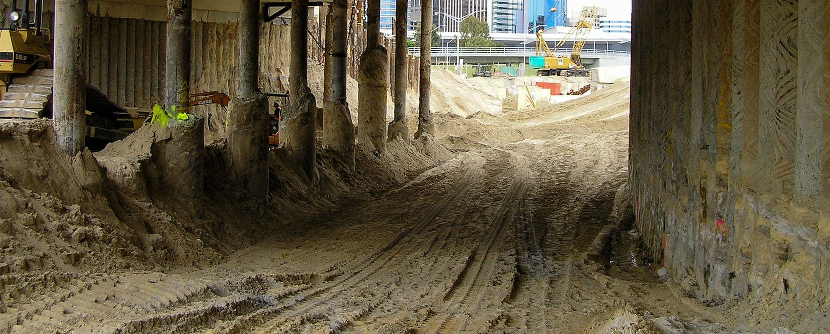 Construction site under a bridge with sandy terrain, heavy machinery and municipal buildings in the background.