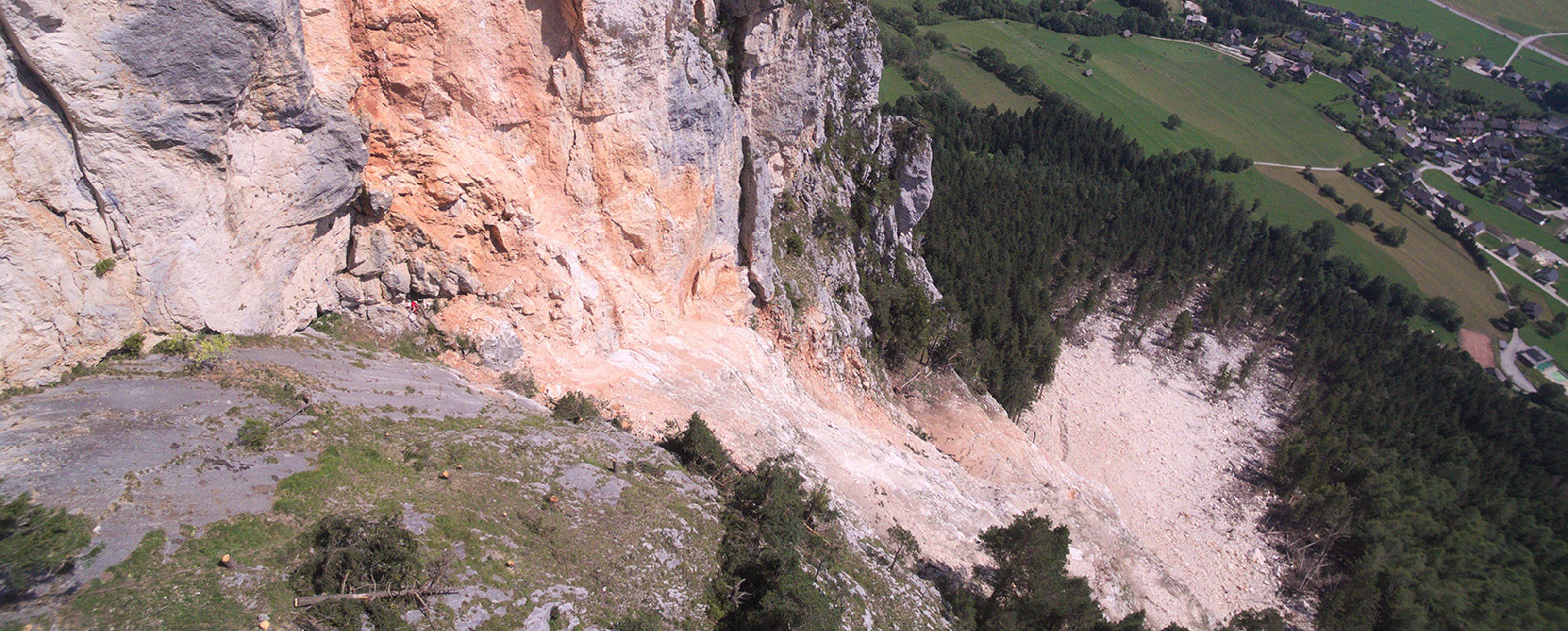Ein Hangrutsch an einer Felswand, umgeben von Wäldern und Wiesen, mit einer Siedlung im Hintergrund.
