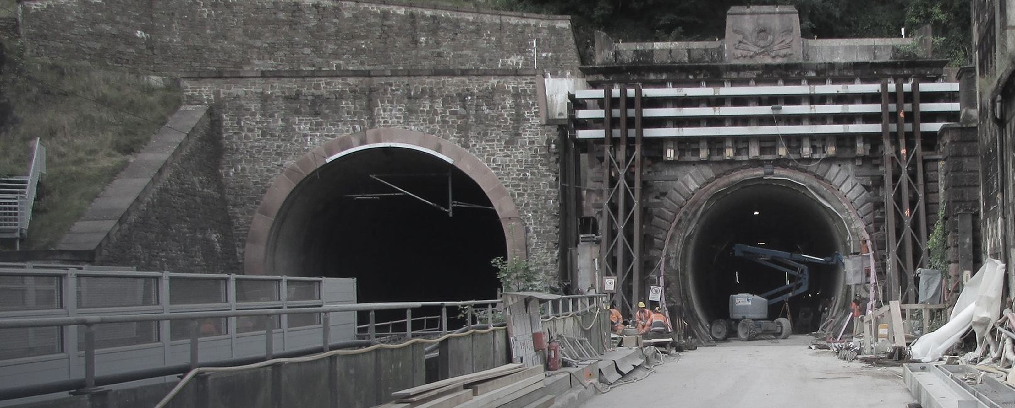 Blick auf zwei Tunnelöffnungen, eine Baustelle im Vordergrund mit Baugeräten und Arbeitern.