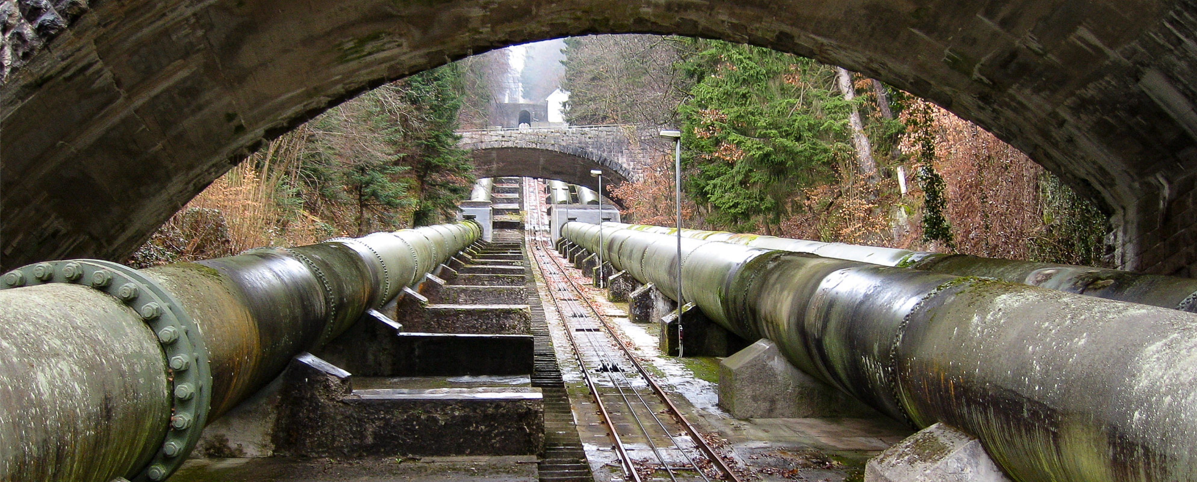 Blick auf große Industrierohre, die unter mehreren Steinbogenbrücken verlaufen, umgeben von Bäumen und moosbewachsenen Strukturen.