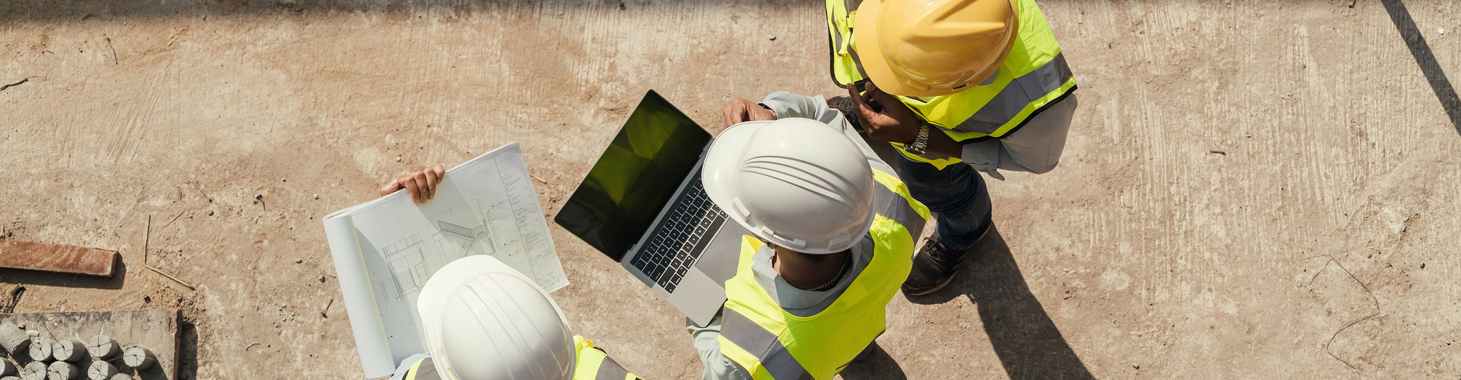 Three construction workers in protective clothing check plans and a laptop on a building site.