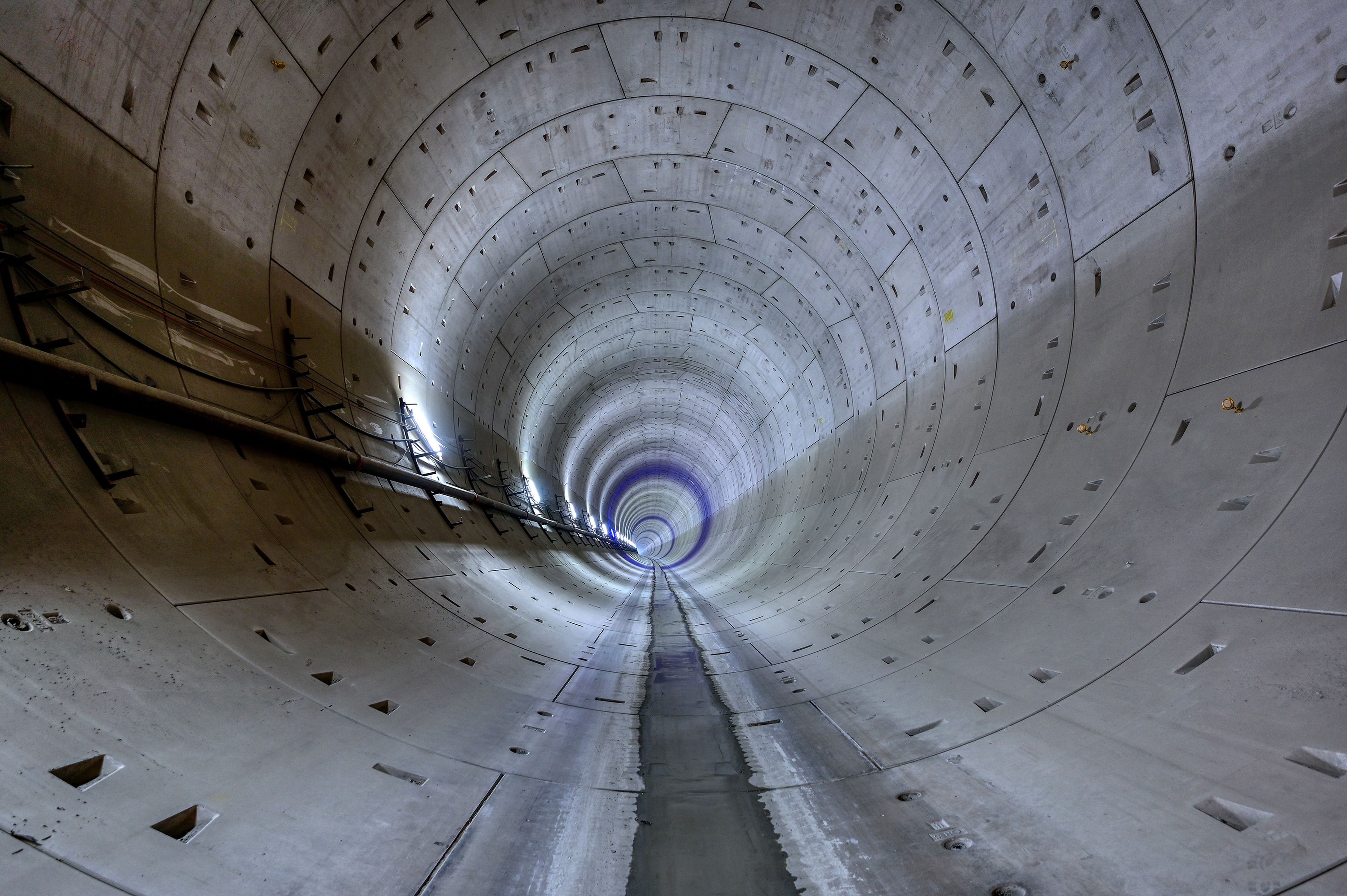 Interior view of a large, circular concrete tunnel with a smooth floor that extends into the distance and is lined with cables on the left wall.
