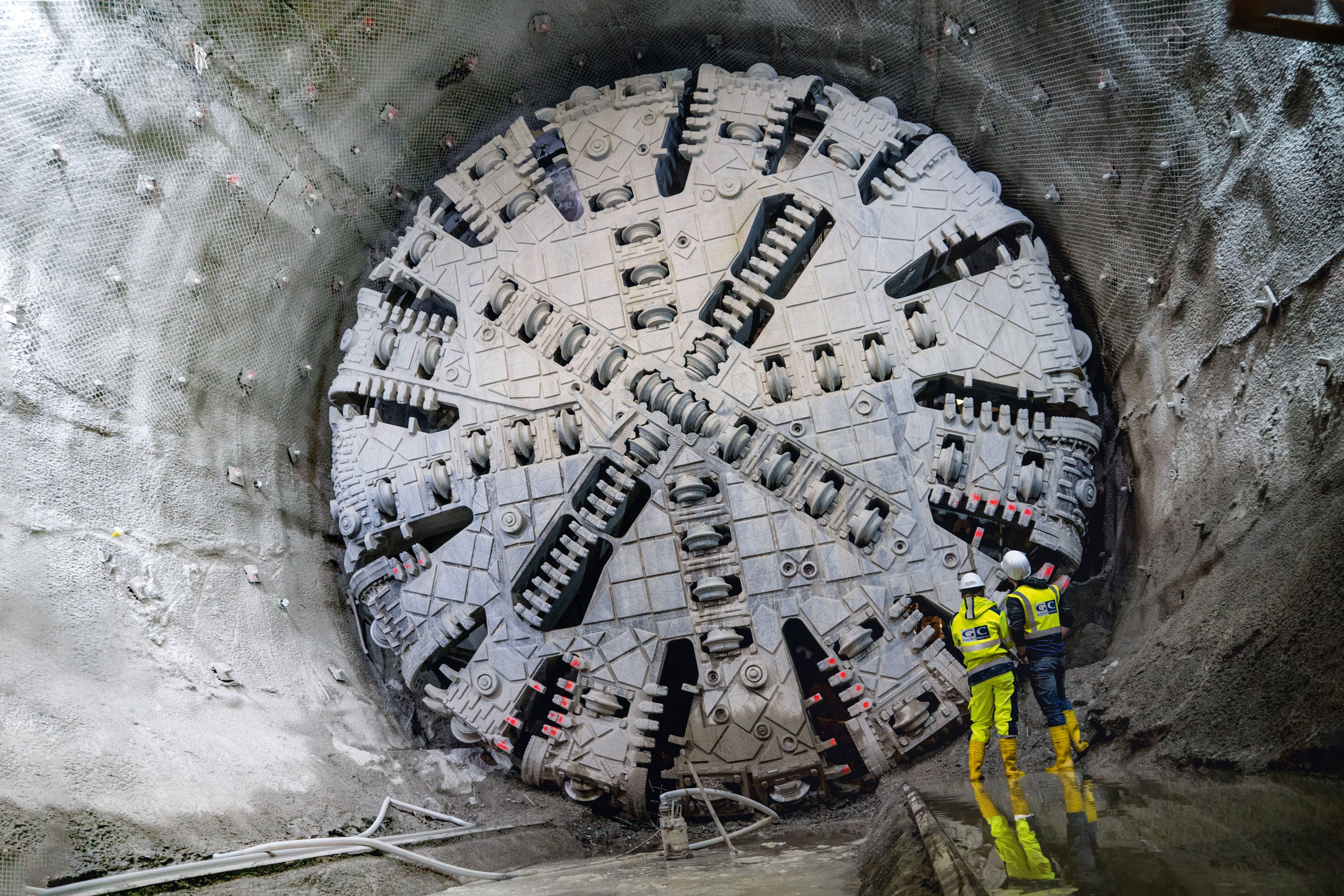 Two workers in protective equipment stand in front of a large tunnel boring machine in a partially excavated tunnel.