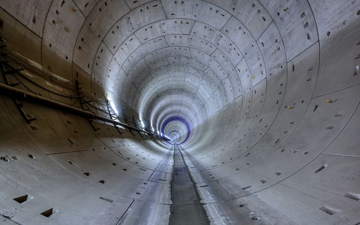 Innenansicht eines großen, kreisförmigen Betontunnels mit glattem Boden, der sich in die Ferne erstreckt und an der linken Wand von Kabeln gesäumt ist.